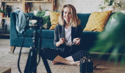 woman with blonde hair sitting on the floor in front of a blue sofa talking to camera demonstrating make up