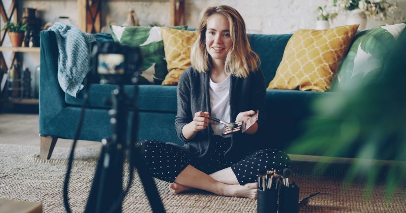 woman with blonde hair sitting on the floor in front of a blue sofa talking to camera demonstrating make up